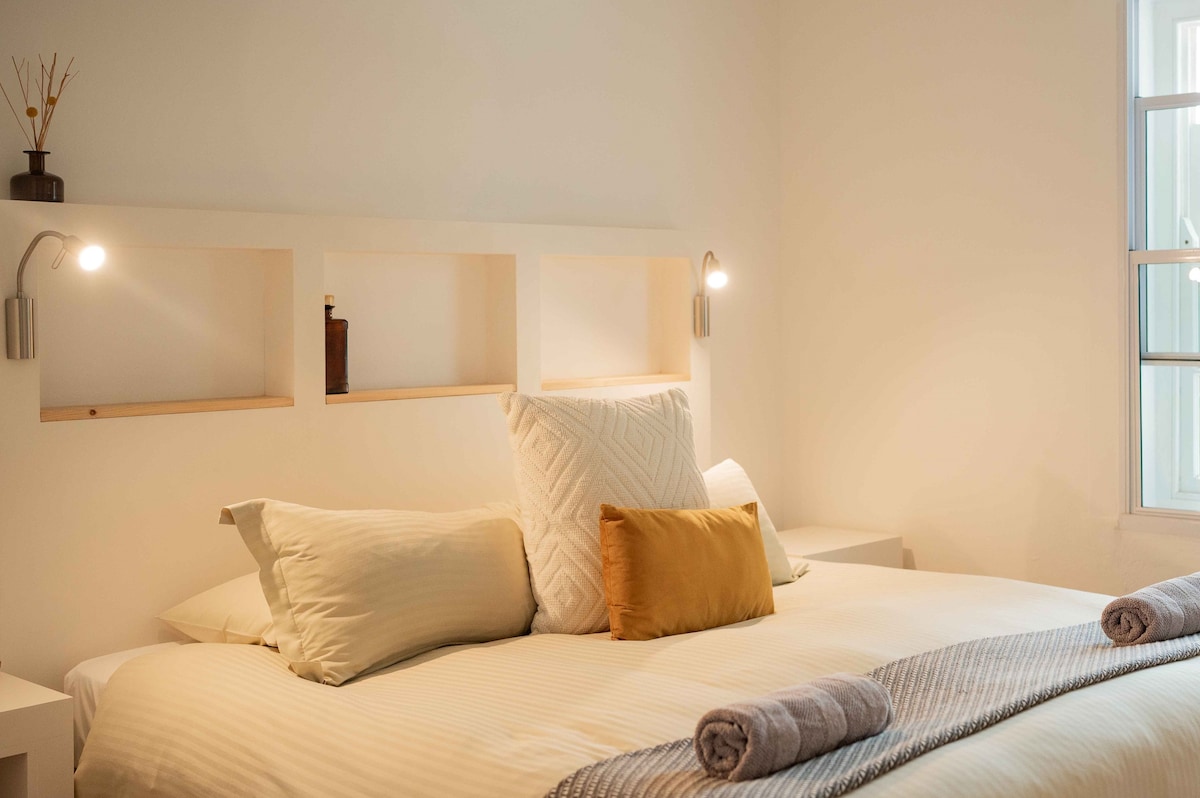 A bright and minimalist bedroom features a spacious bed with soft linen, decorated with an assortment of pillows, including a yellow accent pillow. Side lamps provide gentle lighting, while a window allows natural light to fill the room.