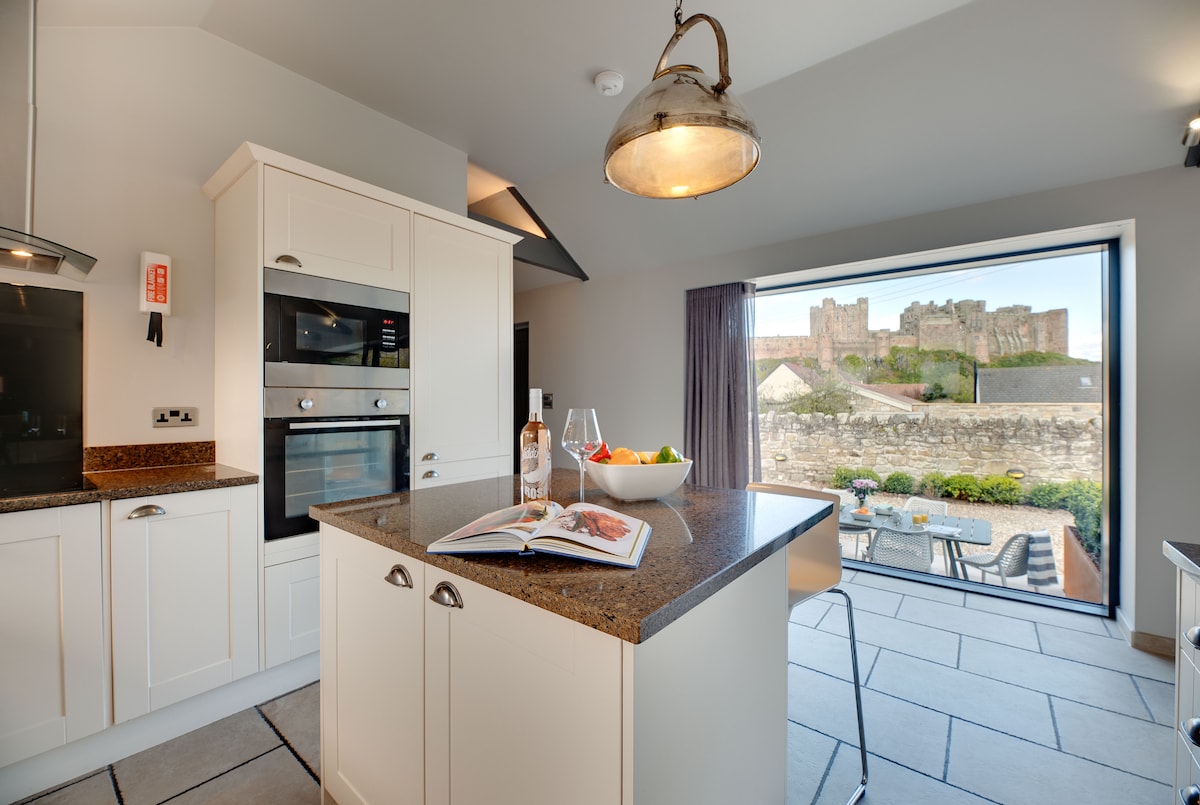 A modern kitchen showcases clean lines and a central island with a textured countertop. A large window offers a panoramic view of Bamburgh Castle and the landscaped outdoor space, while light pours in, enhancing the inviting environment.