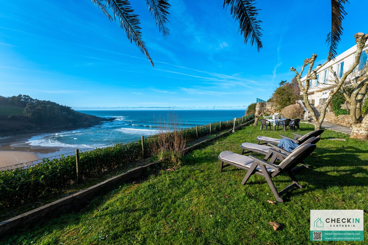 Lounge chairs are positioned on a lush green lawn overlooking a sandy beach and the calm ocean. Palm trees provide some shade, while larger trees dot the area. The bright blue sky and gentle waves create a serene coastal ambiance.
