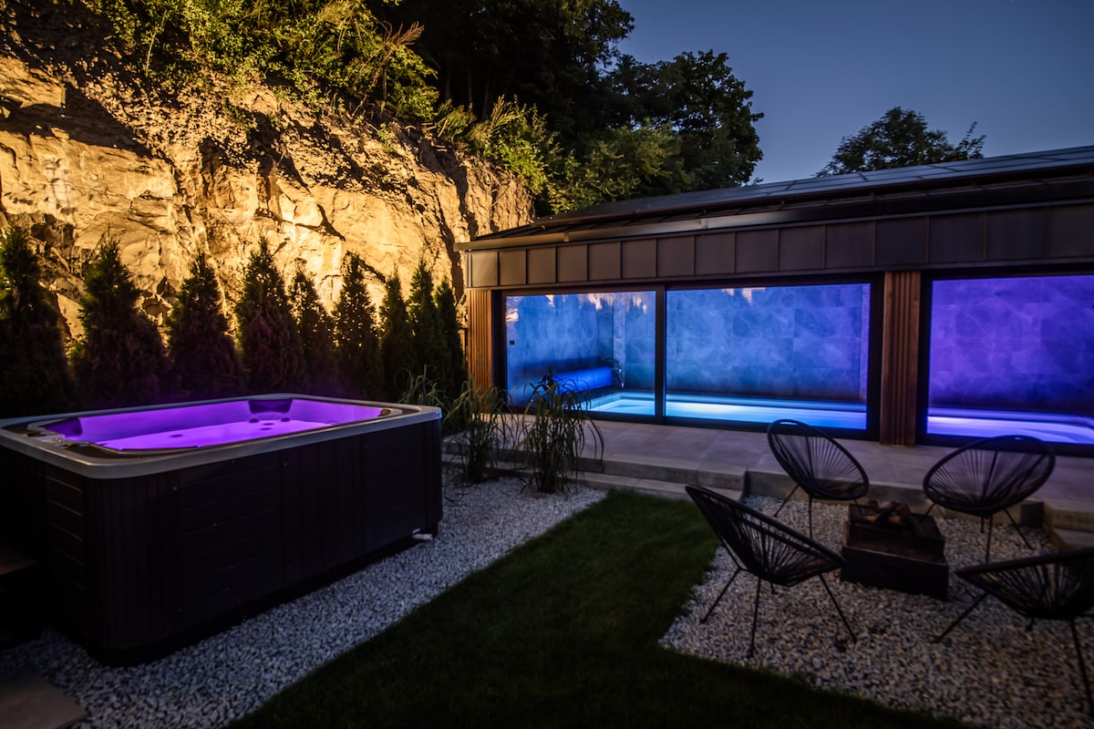An outdoor area is showcased, featuring a hot tub illuminated with purple lighting. Adjacent, a seating arrangement with circular chairs is organized around a small fire pit. A rocky wall is visible in the background, with greenery lining the perimeter.
