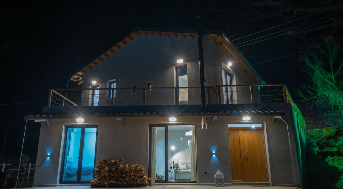 The exterior of a modern lodge is illuminated at night, showcasing a spacious balcony and large glass doors. A stack of firewood is positioned near the entrance, with subtle lighting accentuating the building's features and surrounding greenery.