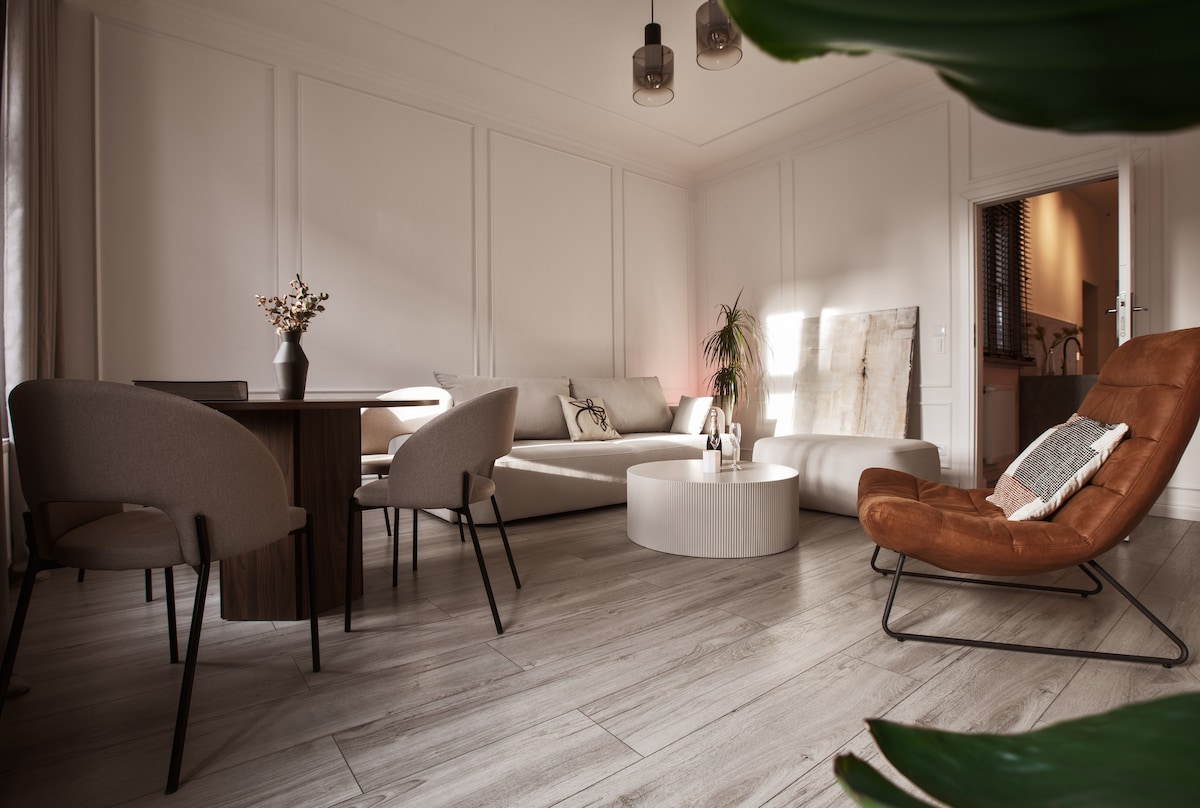 A modern living space features a circular coffee table positioned at the center, surrounded by a comfortable sofa and a pair of armchairs. A dining area includes a dark wooden table with soft, upholstered chairs. Natural light filters in, creating a warm and inviting environment.
