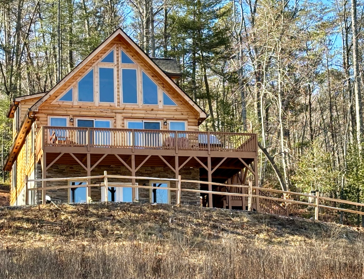 A charming cabin with a wooden exterior is set against a wooded backdrop. Large windows reflect natural light and offer scenic views. A spacious deck, furnished with chairs, extends across the front, providing an inviting outdoor space for relaxation.