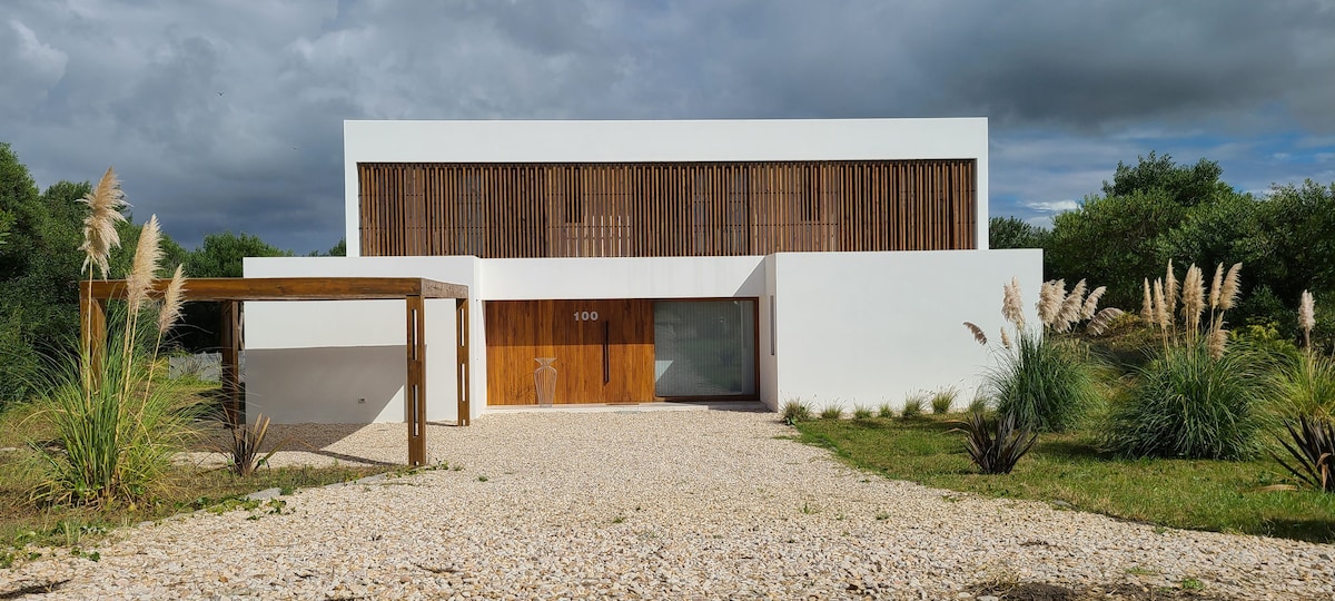 The exterior of the property features a modern white facade with wooden accents, surrounded by a landscaped area. Gravel paths lead to the entrance, which showcases large wooden doors and expansive windows, framed by natural greenery.