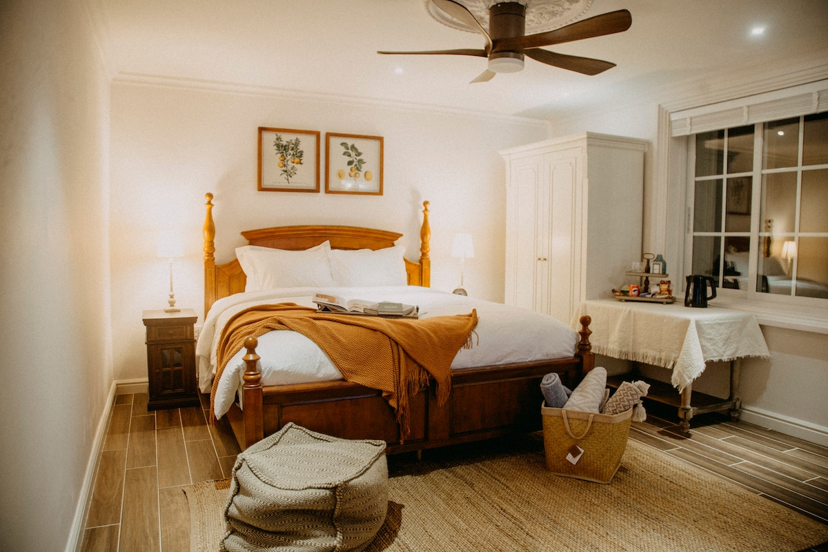 A spacious bedroom features a large wooden bed with decorative pillows and a soft throw draped over the side. Two bedside lamps provide gentle light, while a fan is mounted on the ceiling. A cozy seating area is visible with a small table and a basket on the floor.