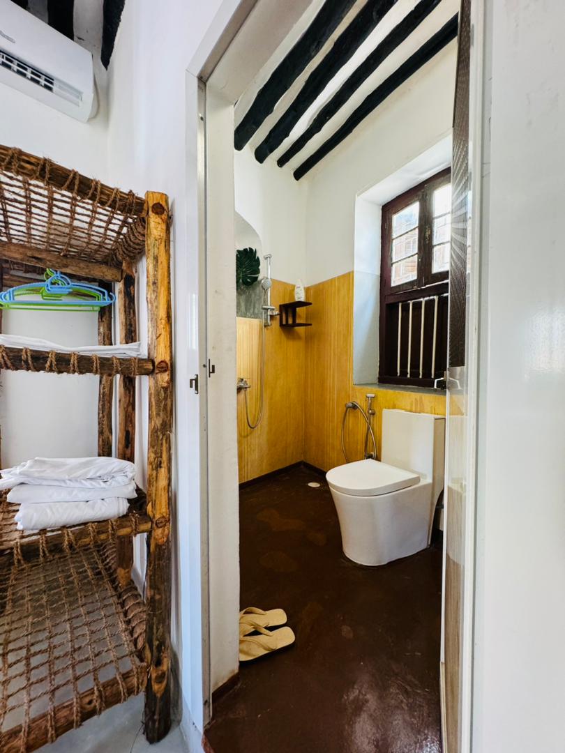 The bathroom features a combination of warm wooden accents and neutral tones. A modern toilet is positioned beside a shower with a handheld fixture. Shelves crafted from natural materials display neatly folded towels and additional amenities, while a window allows natural light to enter the space.