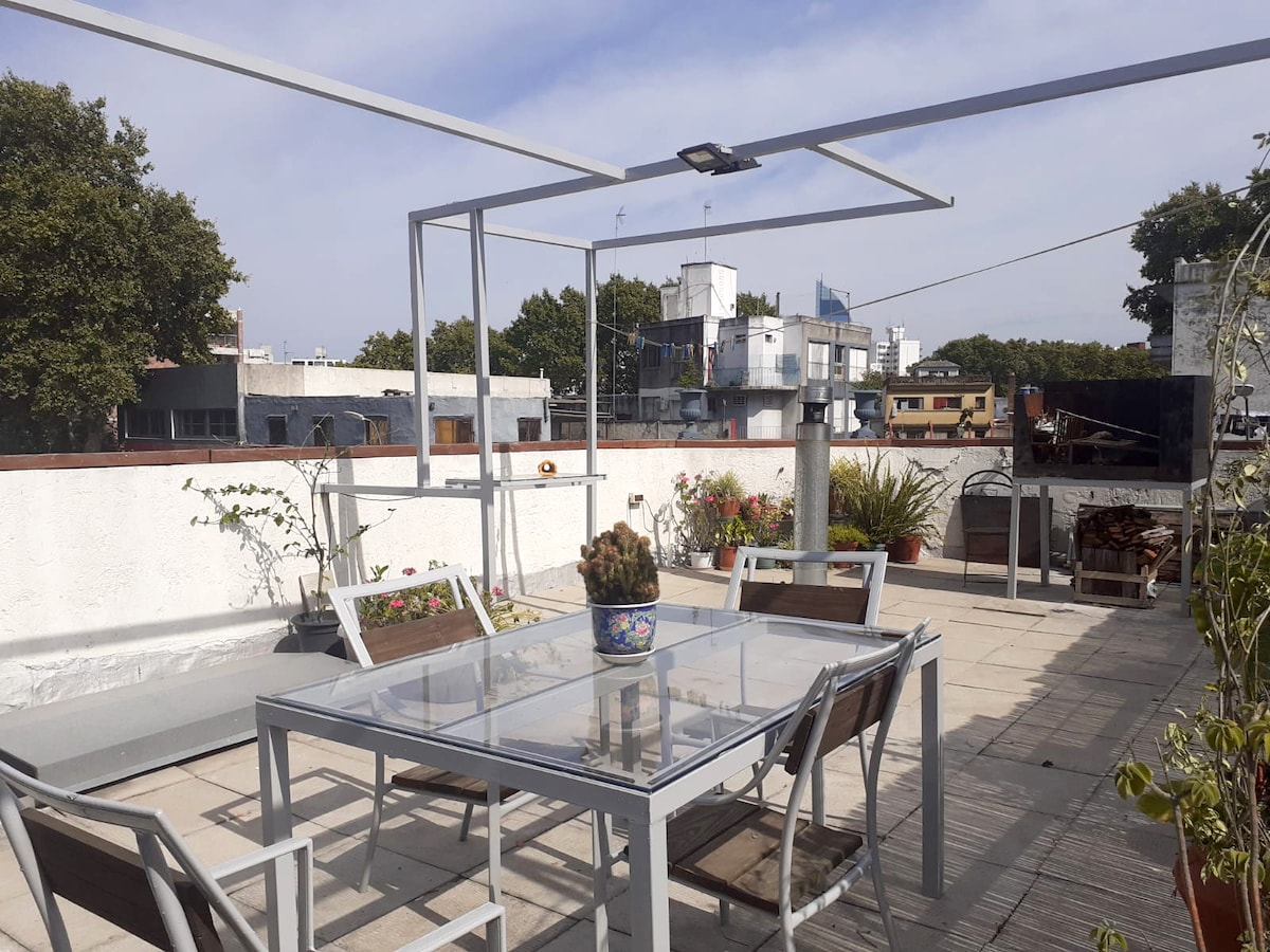 A rooftop terrace features a modern table and chairs, providing ample space for outdoor dining. Potted plants add greenery to the setting, while a barbecue area is visible. The surrounding skyline includes various buildings, enhancing the urban atmosphere.