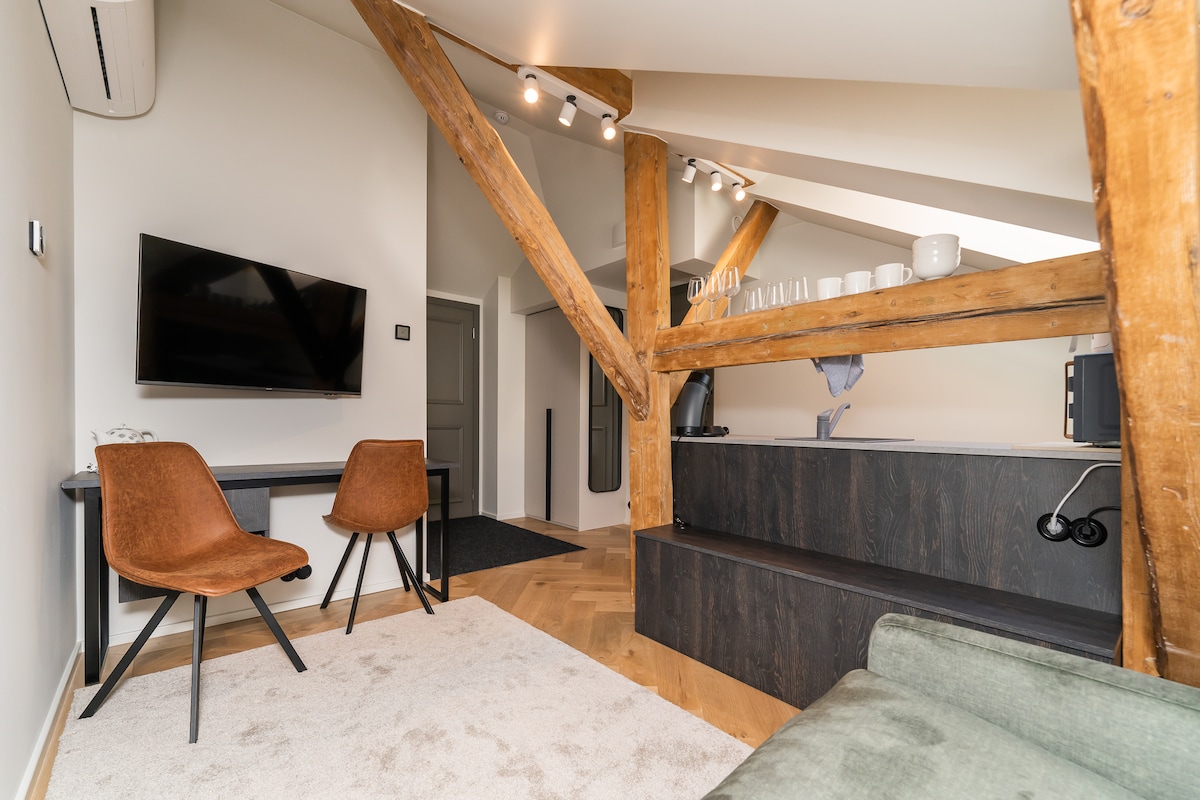 A stylish living area is highlighted by exposed wooden beams and modern furnishings. Two brown chairs and a sleek desk are positioned near a wall-mounted television. A small kitchenette area features dishes arranged on a shelf, enhancing the functional design.