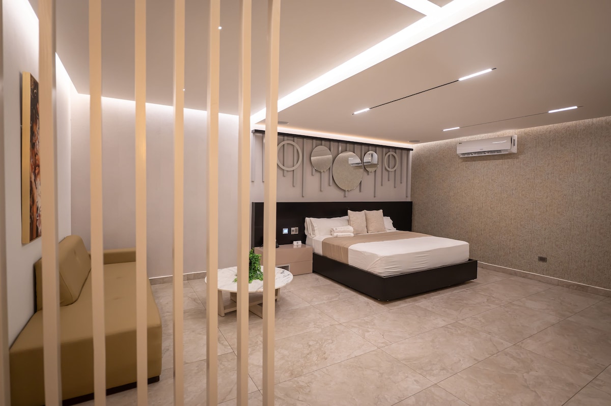 A spacious suite is presented with a modern design, featuring a large bed with neatly arranged bedding. A comfortable seating area is visible, complemented by a round coffee table. Soft lighting highlights the textured walls and unique round mirrors above the bed.