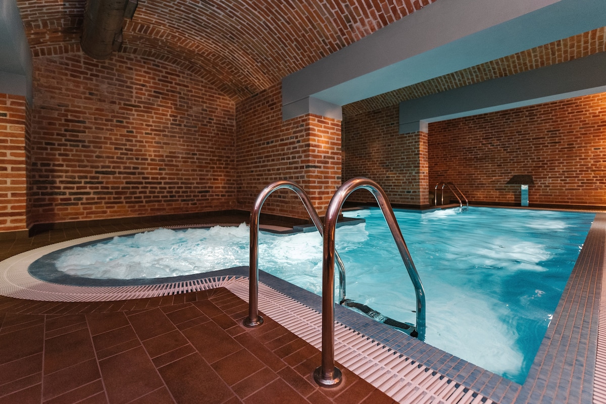 An indoor spa area features a soothing whirlpool surrounded by exposed brick walls. Soft lighting enhances the tranquil atmosphere, while a second pool is visible in the background, offering a serene space for relaxation.