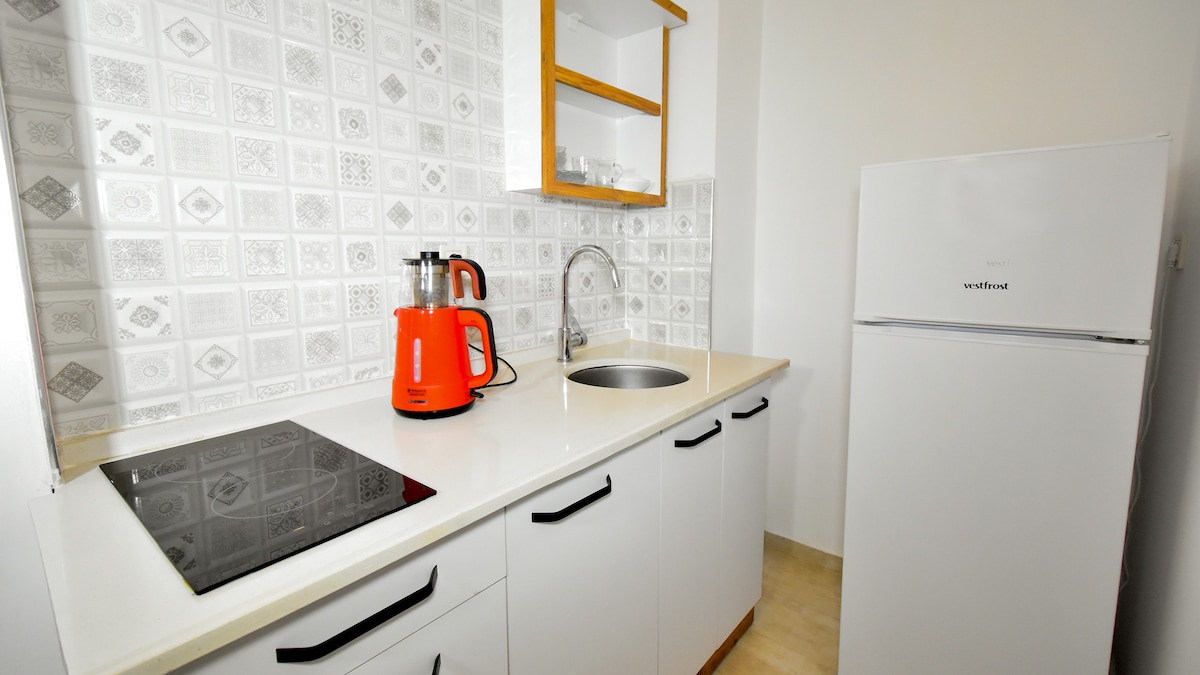 A compact kitchen is presented with modern amenities, featuring a white countertop and cabinets. A refrigerator in the corner is complemented by an orange kettle and a water boiler located beside a small sink. A stovetop is positioned on the surface, enhancing functionality.