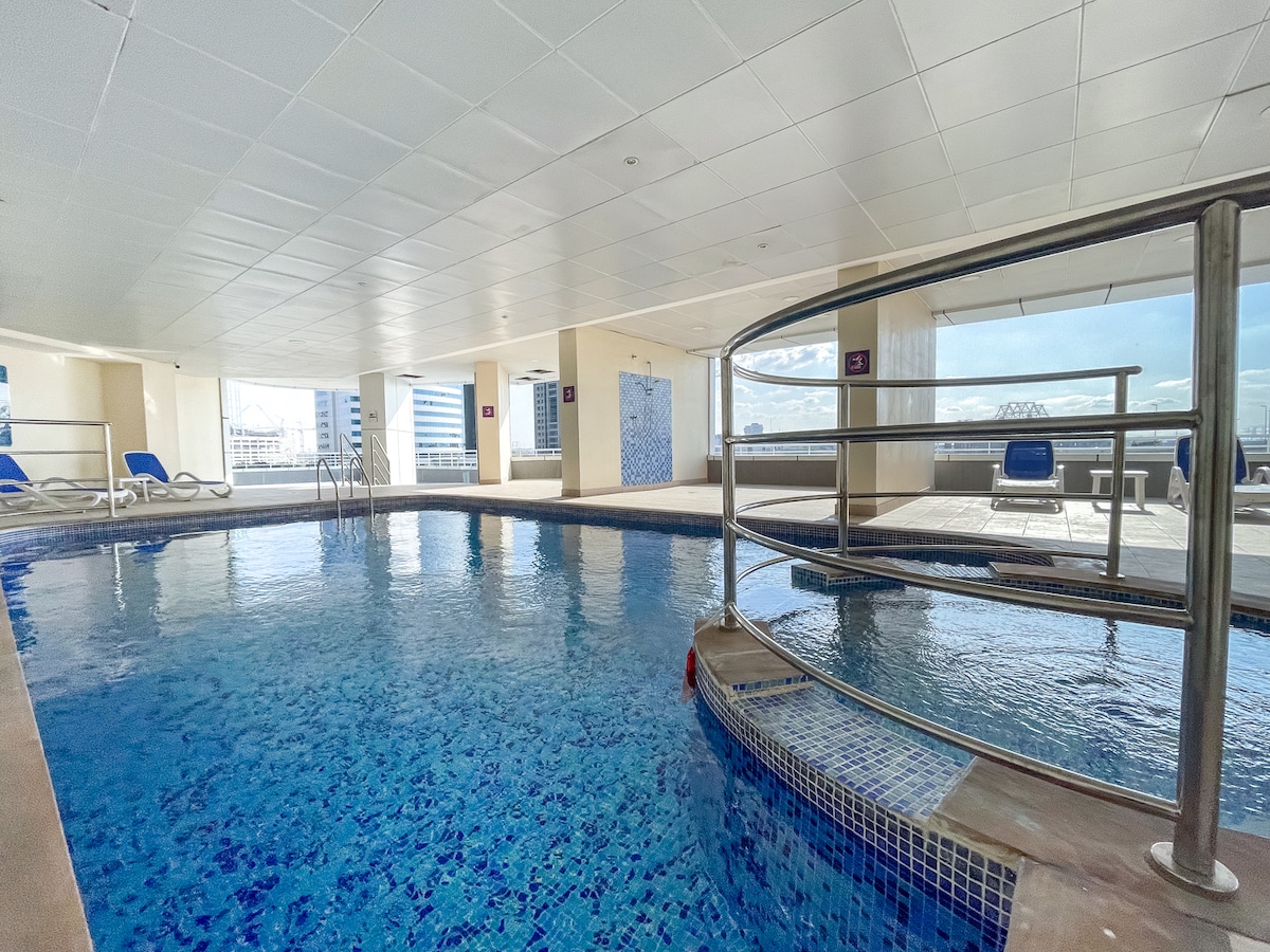 A spacious indoor pool area showcases clear blue water, complemented by surrounding lounge chairs. Natural light floods the space through large windows, creating a bright and inviting atmosphere. A sleek railing encircles a section of the pool, enhancing safety and accessibility.