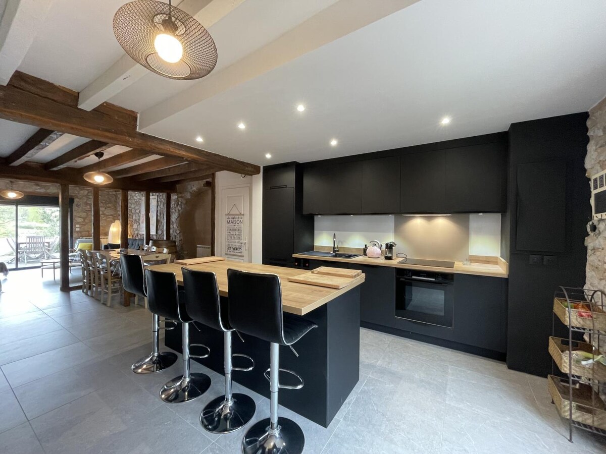 A renovated kitchen is showcased, featuring a central island with wooden countertops and four black bar stools. Modern appliances include an oven and stovetop, while warm lighting complements the natural stone and wooden beams present in the open space.