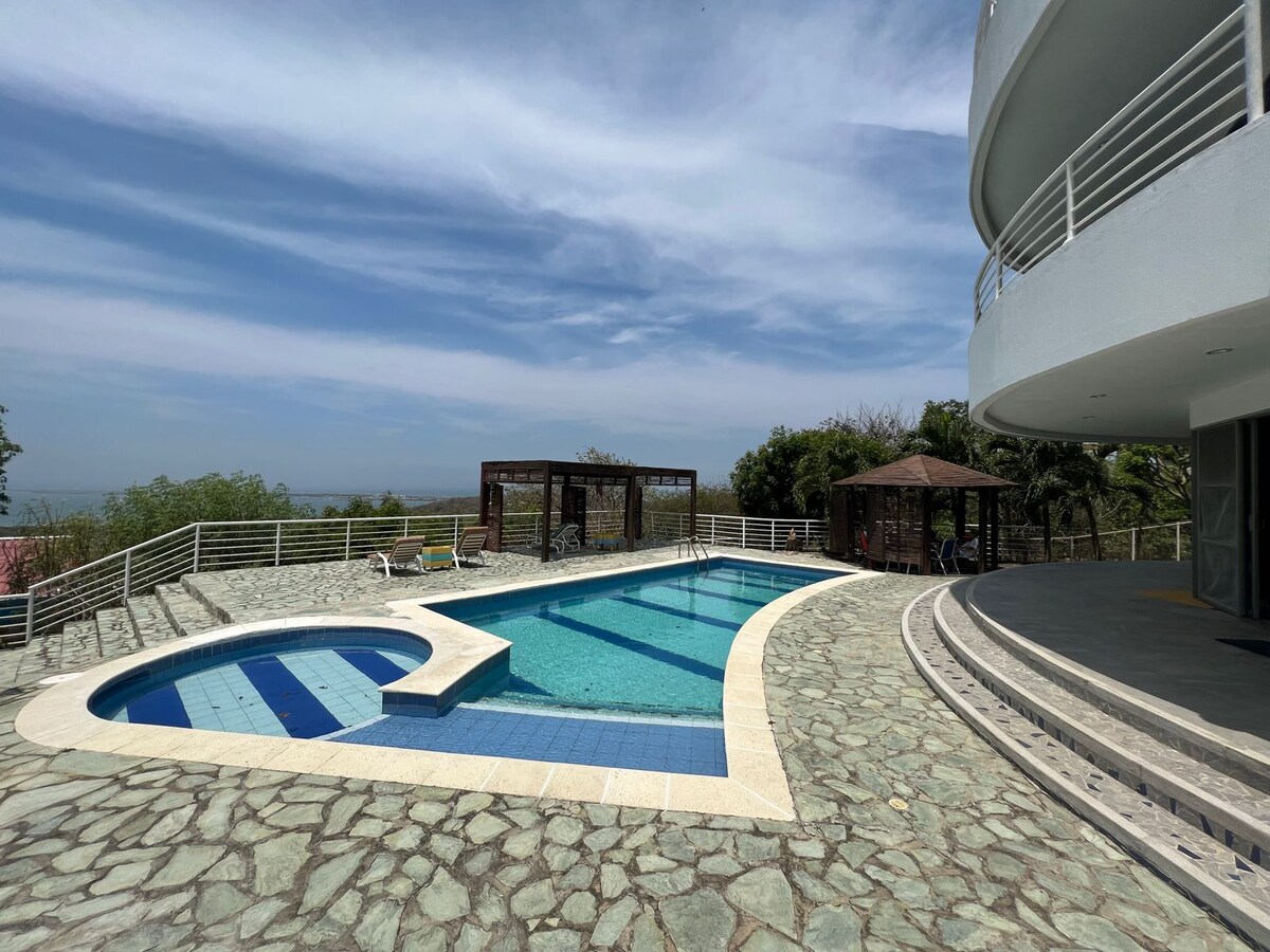 A curved pool, surrounded by a patterned stone deck, reflects the sky. Lounge chairs are positioned near the water's edge, and a shaded gazebo is visible in the background. Lush greenery frames the area, contributing to a serene outdoor setting.