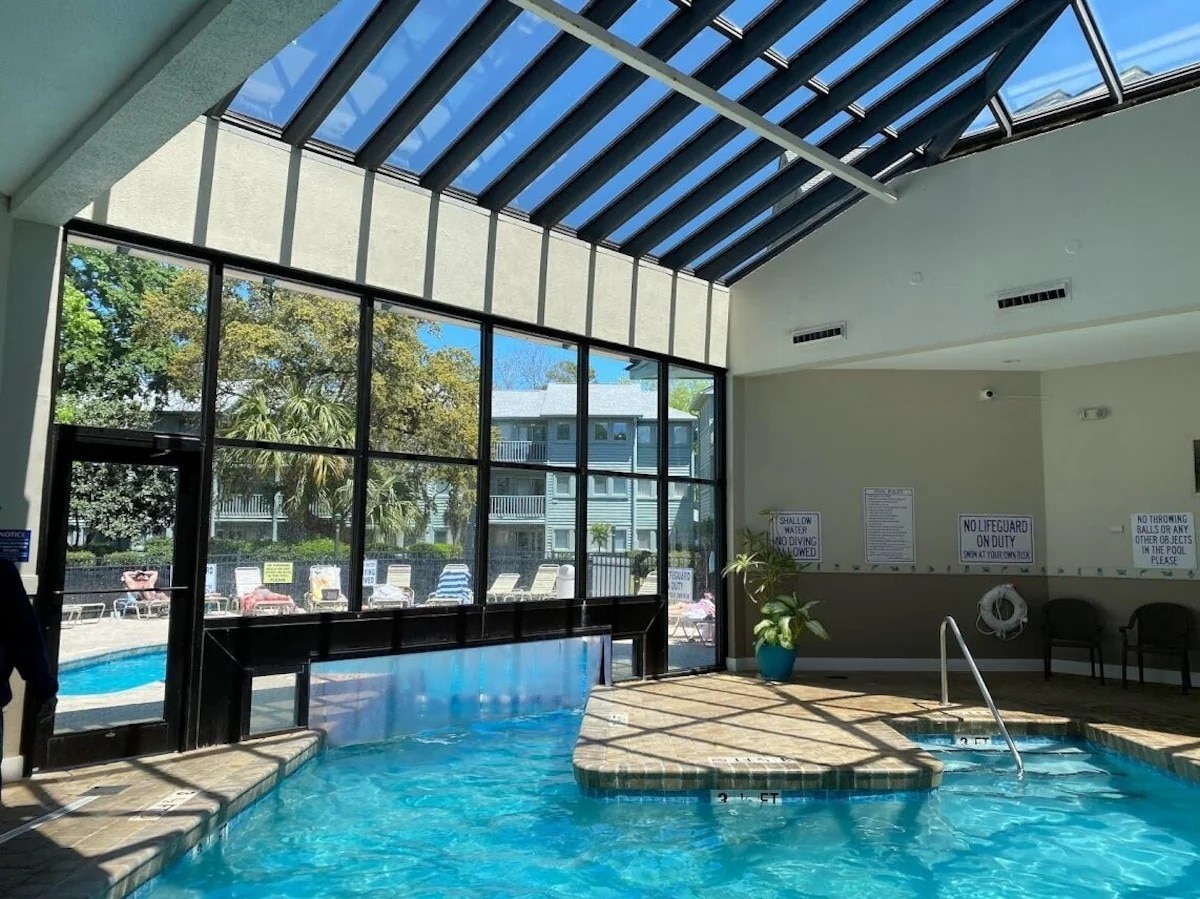 An indoor pool area is highlighted by a glass ceiling, allowing natural light to fill the space. The pool features clear, inviting water with a shallow entry. Lounge chairs line the edge, providing relaxation options. Tropical plants are placed for a touch of nature.