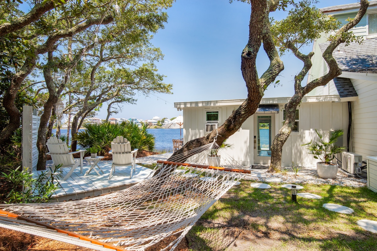 A hammock is suspended between two trees, providing a serene resting spot near the lake. Adirondack chairs are positioned on a deck, surrounded by lush greenery. The calm water reflects the clear blue sky, creating a peaceful atmosphere for relaxation.