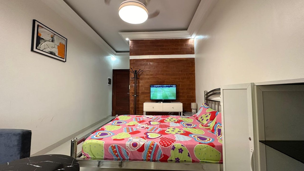 A colorful sleeping area features a spacious bed adorned with vibrant patterned bedding. A flat-screen television is mounted on a wall opposite the bed, with a white media console positioned below. The room is illuminated by a modern ceiling fixture and has a minimalist decor.
