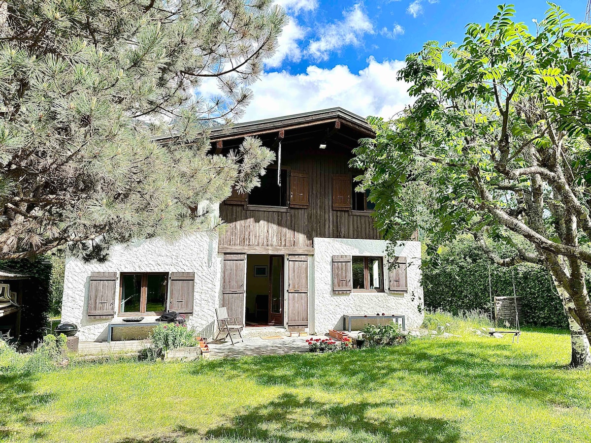 The chalet is surrounded by greenery, showcasing an inviting exterior with wooden accents. Large windows and patio doors allow natural light to fill the space. A grassy area is present, enhanced by trees and shrubs, creating a serene outdoor setting.