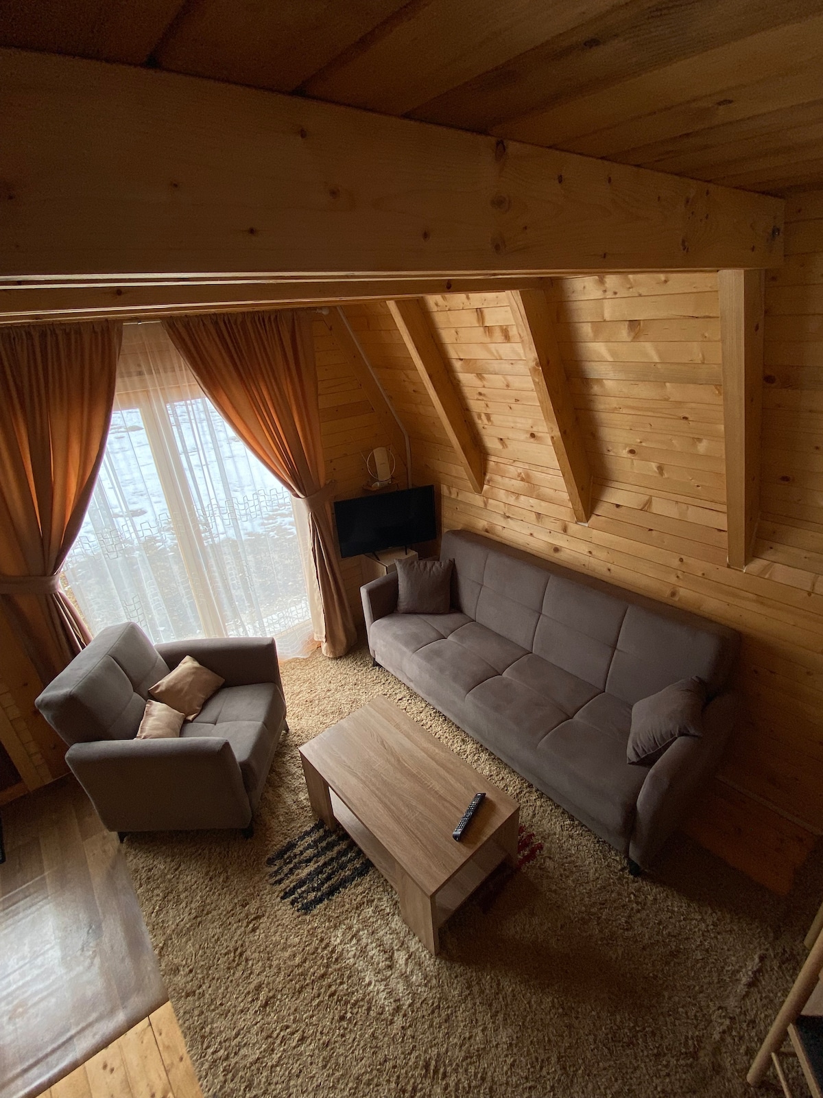 A cozy living area is highlighted by wooden beams overhead and soft, warm lighting. A two-seater sofa and two armchairs are arranged around a central coffee table. Light drapes frame the windows, allowing natural light to fill the space, which features a plush area rug.