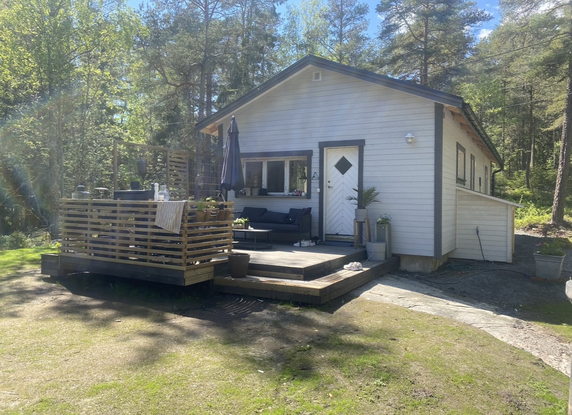 A charming white cottage is surrounded by tall trees, featuring a wooden deck with outdoor seating and a shaded umbrella. The front door, adorned with a diamond-shaped window, opens into a well-lit interior, with a small planter by the side enhancing the entryway.