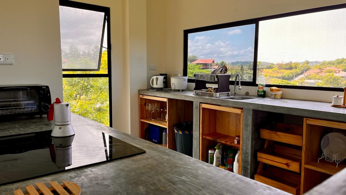 A modern kitchen features concrete countertops and wooden cabinetry. A stovetop is visible alongside essential appliances, including a kettle and toaster. Large windows provide natural light and views of the surrounding landscape, contributing to a serene atmosphere.