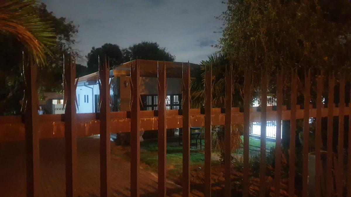 A view of a dwelling surrounded by greenery is captured at night. The structure is partially obscured by a decorative fence, revealing large windows that reflect soft outdoor lighting. The landscape features well-maintained grass and several trees in the vicinity.