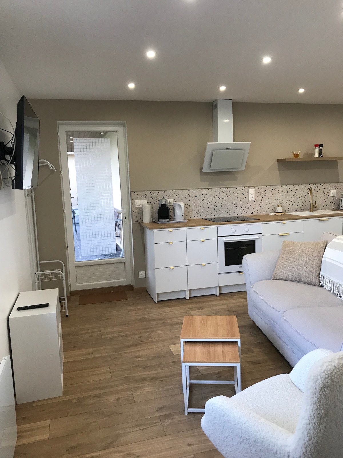 A welcoming living area features a comfortable sofa and a compact coffee table. The kitchen has modern appliances, including a microwave and an oven, set against a versatile backsplash. Natural light enters through a nearby door leading to an outdoor space.