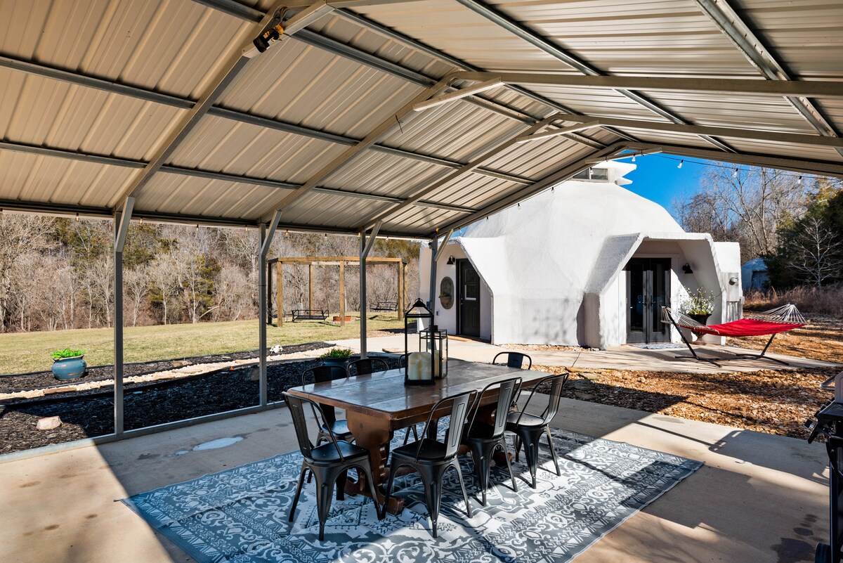 An outdoor covered patio features a large wooden dining table surrounded by multiple metal chairs. A patterned rug lies beneath the table, with a hammock and swings visible beyond the dining area, all set against a backdrop of natural greenery and the Dome structure.