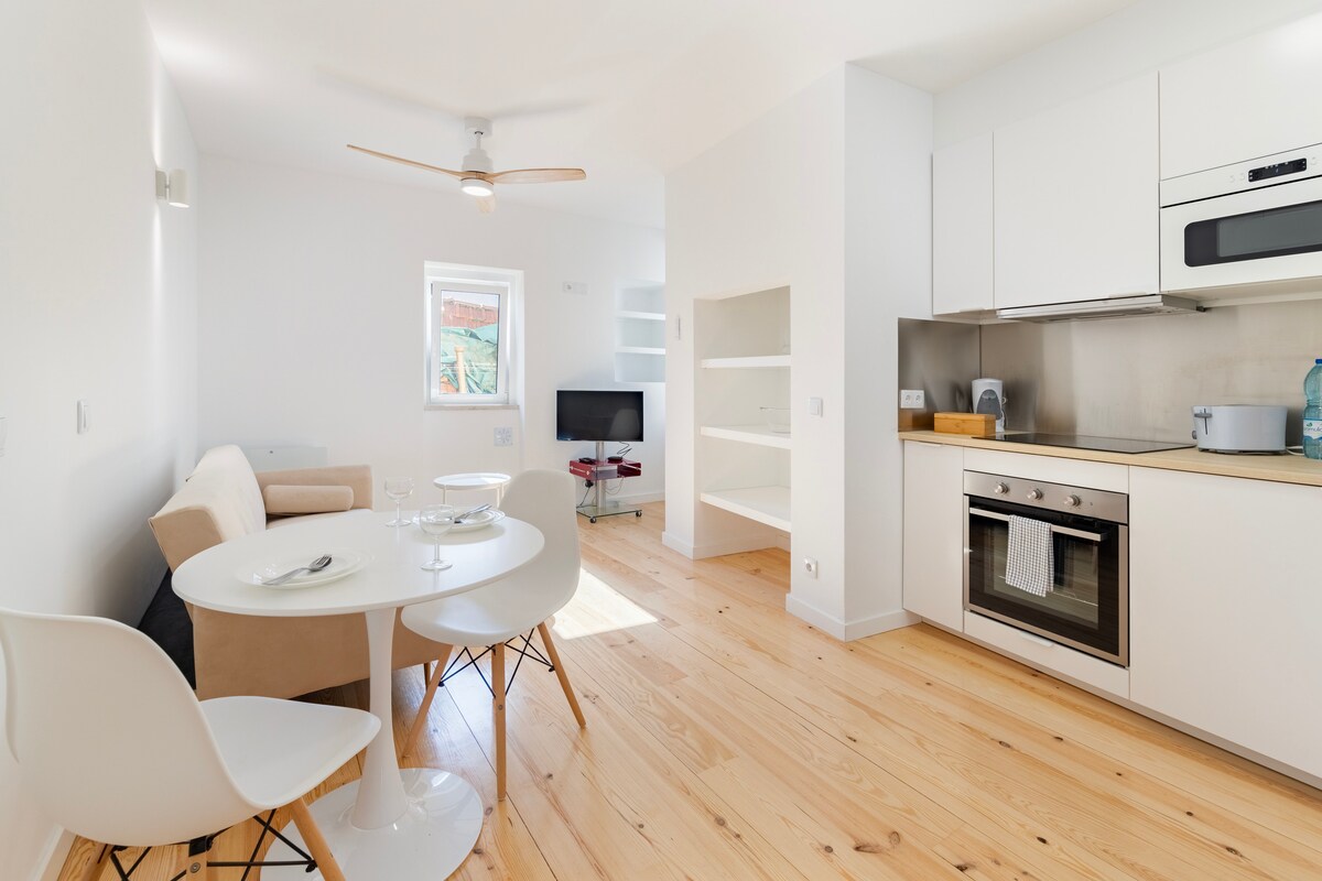 A modern, open-plan living space is featured, showcasing a circular dining table paired with white chairs. A cozy sofa is positioned nearby, with a TV mounted on the wall. Natural light floods the room through a window, highlighting the warm wooden flooring.