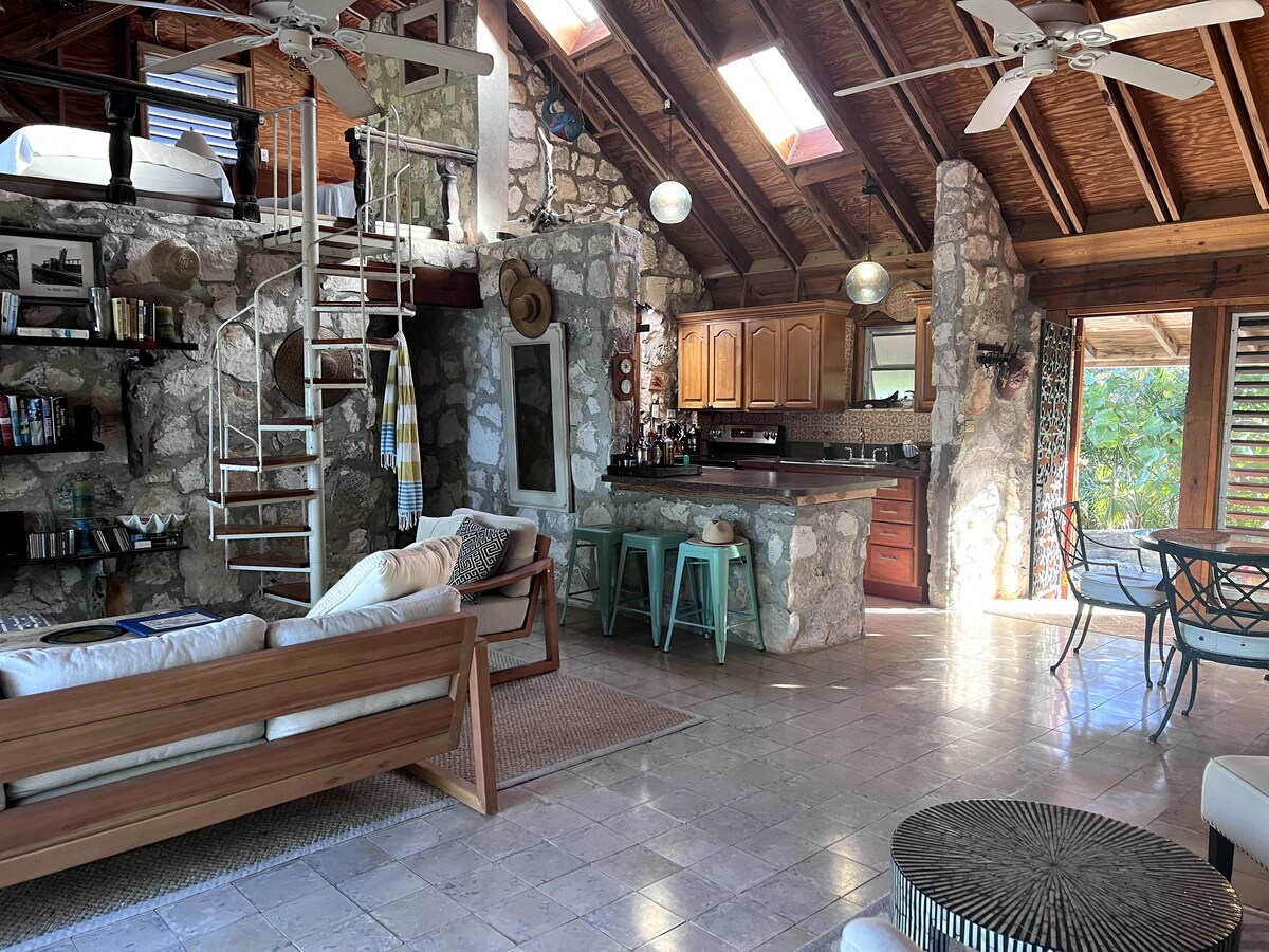 An inviting open-plan living area features natural stone walls and high wooden ceilings. A spiral staircase leads to an upper loft. The kitchen is equipped with wooden cabinetry and metallic bar stools. A cozy seating arrangement includes a light-colored sofa, complemented by decorative accents.