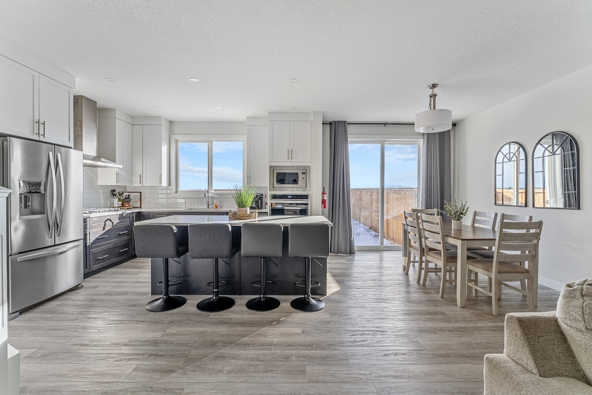 An open-concept kitchen and dining area features a modern kitchen with stainless steel appliances and a large island with four black bar stools. A dining table with chairs is positioned near large windows. Natural light fills the space, highlighting the smooth wooden flooring.