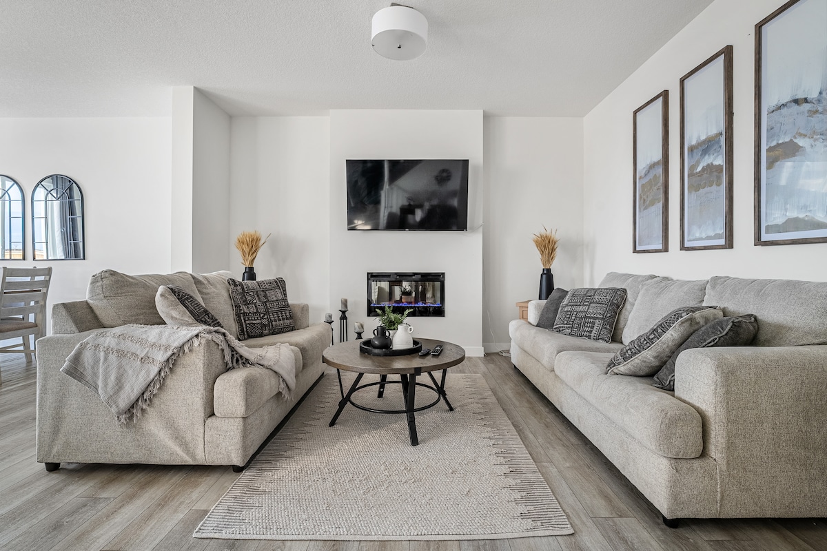 A spacious living room features two light-colored sofas positioned around a circular coffee table. A minimal area rug anchors the seating area. A television is mounted on the wall above a modern fireplace, with decorative wall art and plants adding a touch of elegance.
