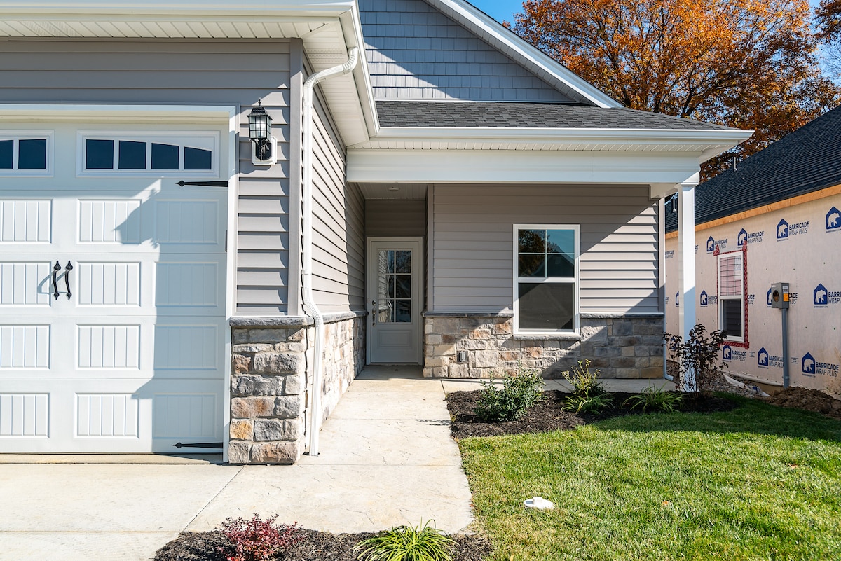 The exterior of the cottage features a combination of stone and siding, with a welcoming entry area. A driveway leads to a garage door, and landscaped gardens with plants border the path, enhancing the approachable entrance.