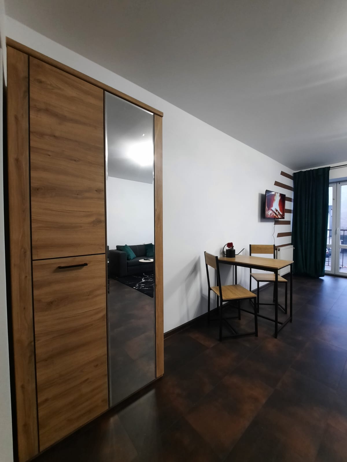 A wooden wardrobe with a mirror stands against a white wall. Nearby, a small dining set features two wooden chairs and a black metal table. A colorful artwork is displayed on the wall, and a green curtain frames a nearby window, leading to an outdoor view.