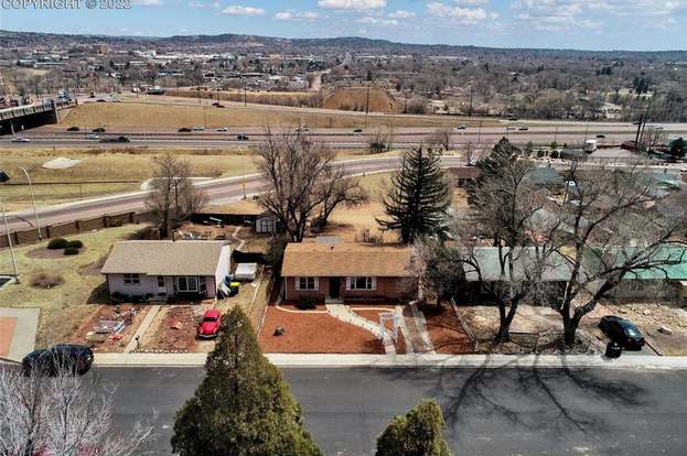 The neighborhood is presented from an aerial view, showcasing single-story houses with landscaped yards. Nearby roads and open fields are visible, along with distant mountain ranges under a clear sky. The layout of the area includes well-maintained streets and ample green spaces.
