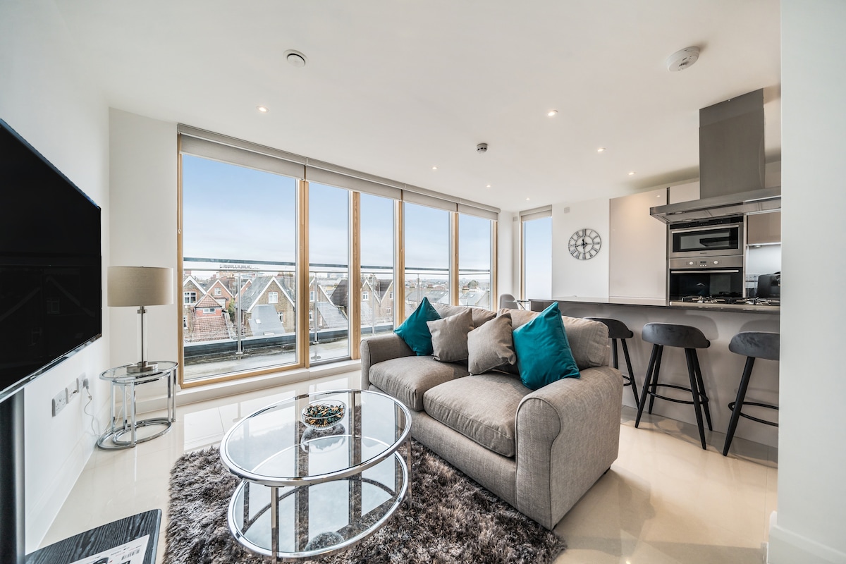 A spacious living area is showcased, featuring a comfortable gray sofa adorned with turquoise cushions. Natural light floods through large glass windows, providing views of the surrounding area. A round glass coffee table sits on a soft rug, accompanied by high stools near the kitchen.