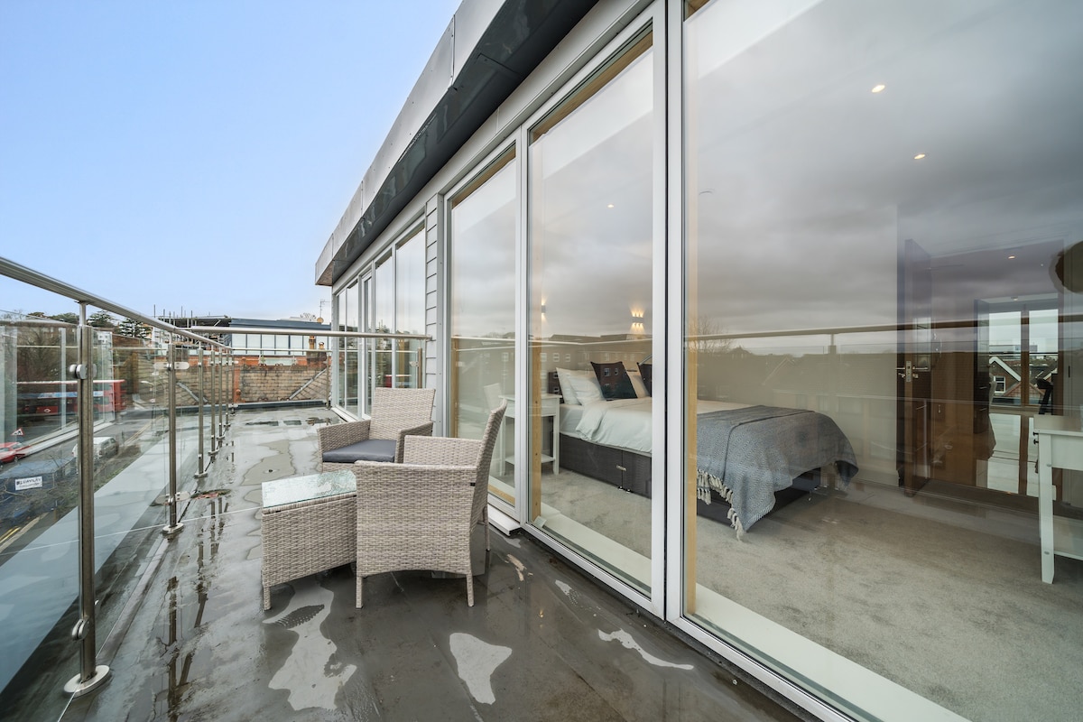 A private balcony is shown featuring two woven chairs positioned around a small table. Expansive glass doors offer a view of the interior space, revealing a neatly made bed with a soft blanket. The balcony provides a clear perspective of the surrounding area.