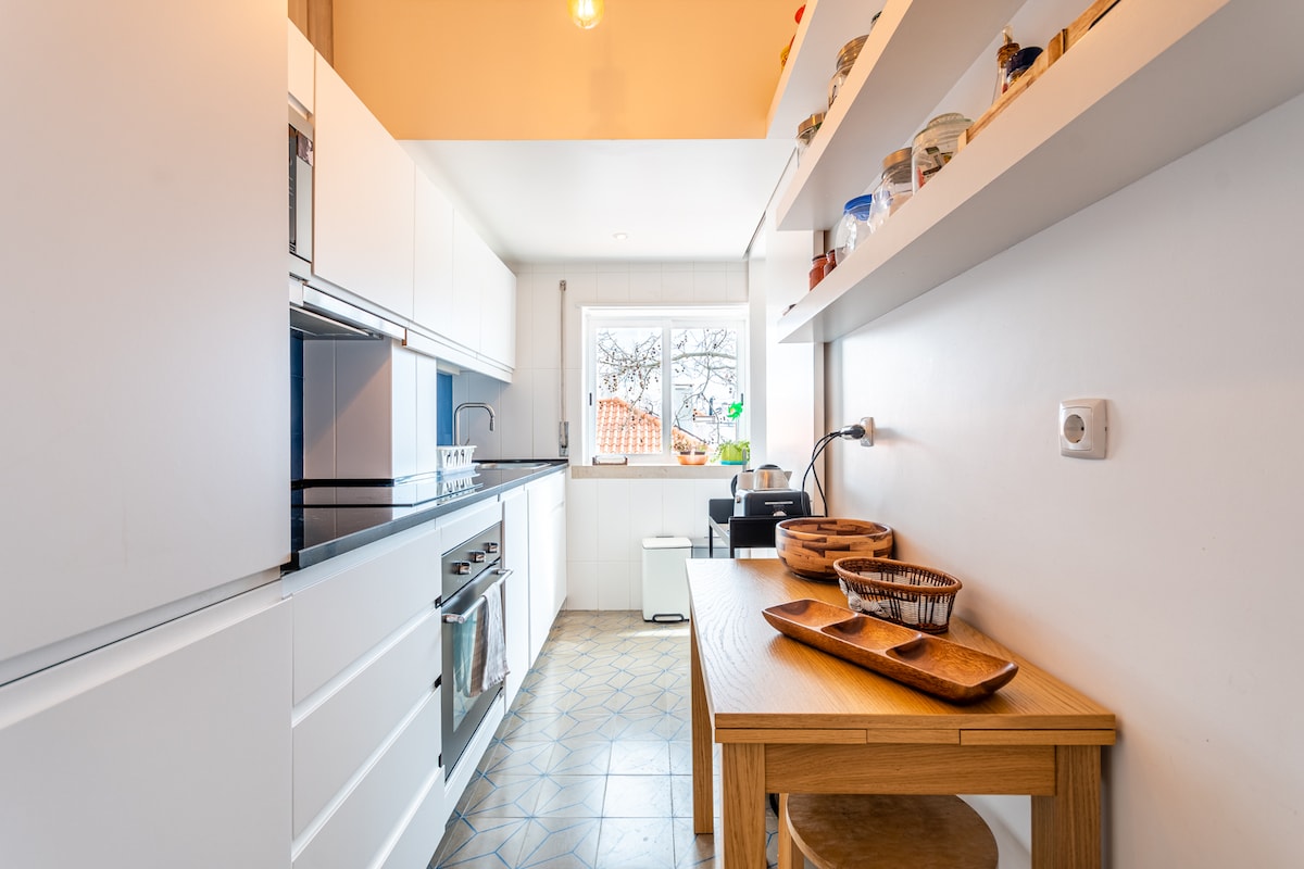 A modern kitchen is shown, featuring sleek white cabinetry and a spacious countertop with wooden serving trays. Natural light enters through a window, illuminating the area. A coffee machine and additional kitchen storage are visible, contributing to the functional layout.