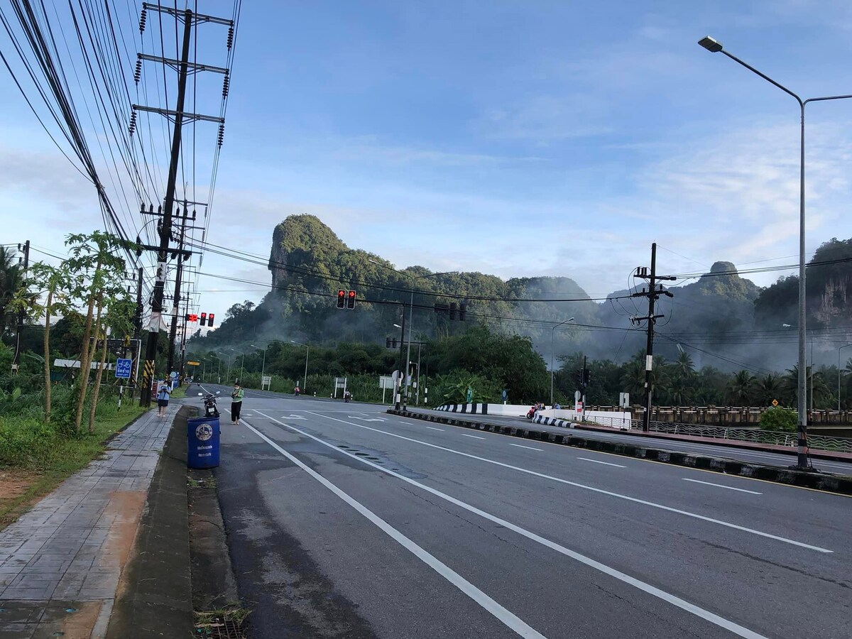 A serene view of a wide road, bordered by lush greenery and mountains in the background. Mist gently rises from the ground, creating a tranquil atmosphere. Traffic lights and utility poles are visible, indicating a well-maintained area. The sky is partially cloudy, enhancing the calm scenery.