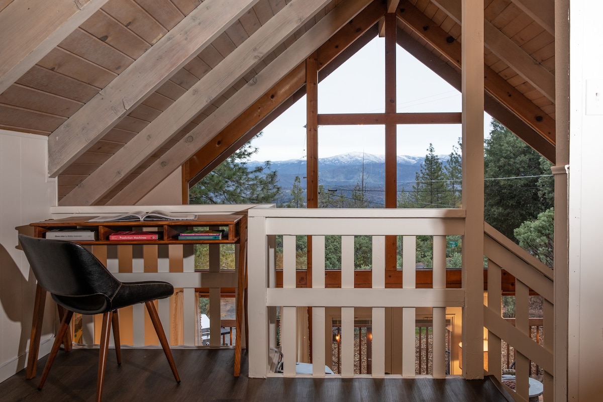 A bright loft area features a stylish chair next to a desk, overlooking large windows that frame captivating views of the surrounding mountains and trees. Wooden beams and warm-toned walls create a cozy ambiance.