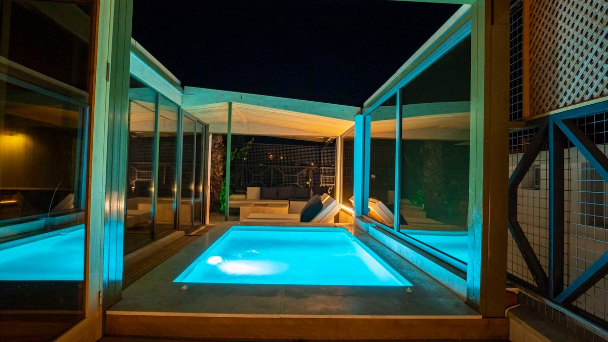 A tranquil pool area is illuminated in soft turquoise light, surrounded by expansive glass walls that create an open feel. Comfortable seating is arranged nearby, inviting relaxation beneath the night sky.