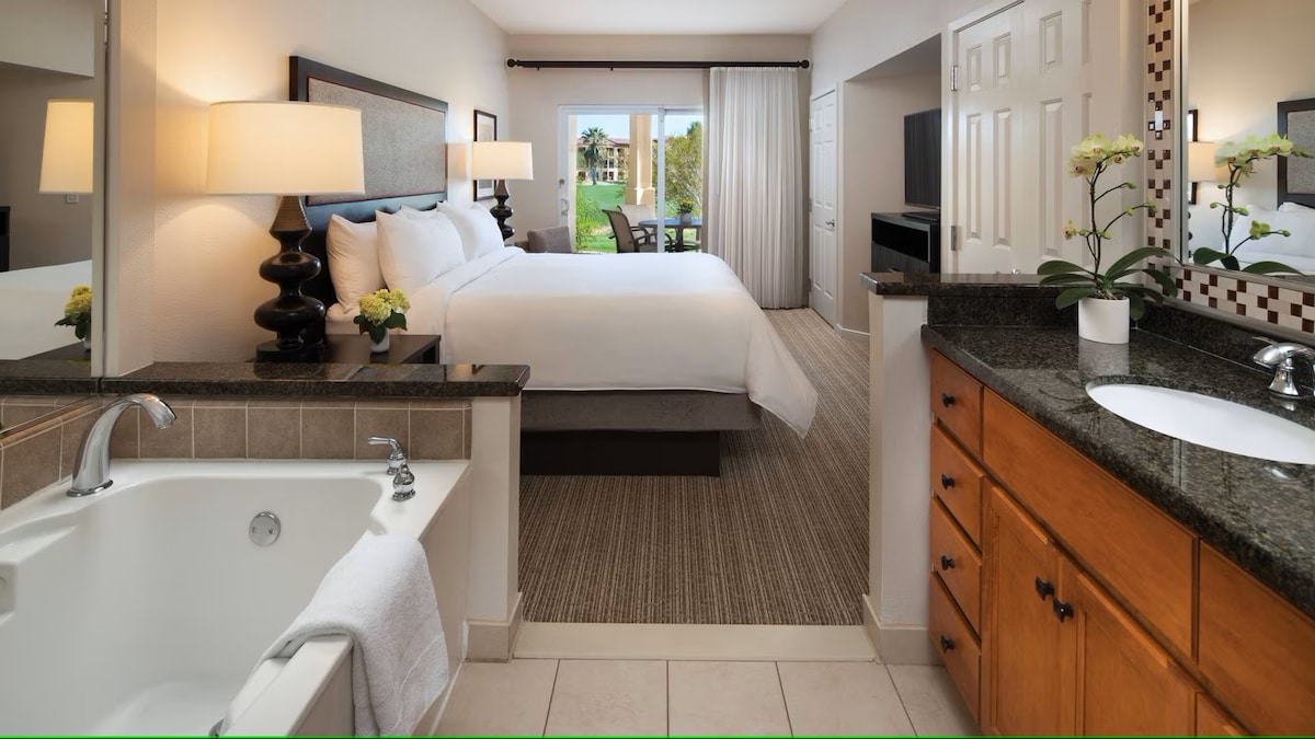 A spacious bathroom area features a soaking tub beside a double vanity with a granite countertop. The adjoining area includes a large bed and a seating space with large windows allowing natural light and views of the outdoors.