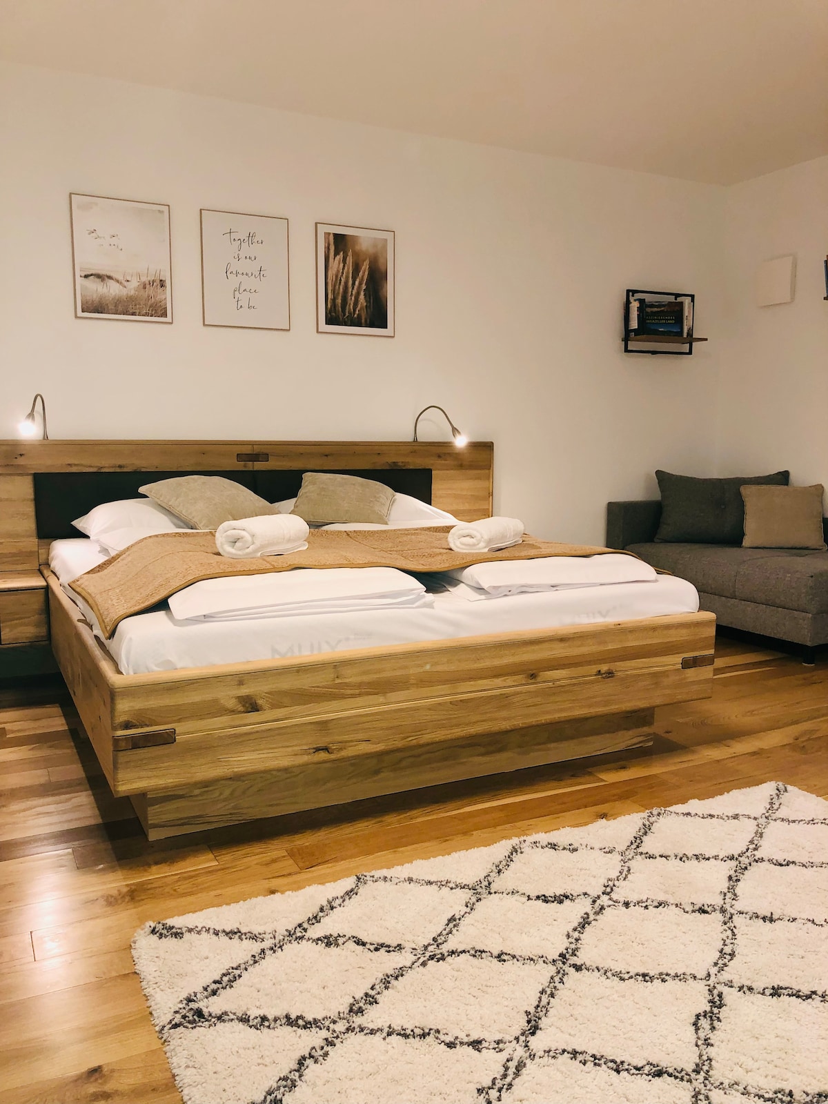 A modern sleeping area features a spacious wooden bed with cozy white linens and decorative pillows. Two bedside lamps are positioned above the bed, illuminating art on the wall. A comfortable sofa and a soft area rug add warmth to the room.
