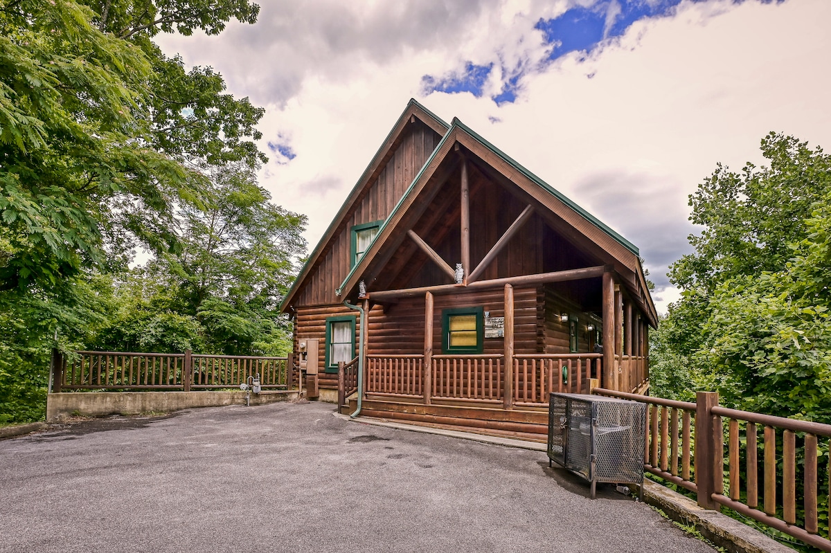 The exterior of a modern cabin is shown, featuring a large gabled roof and wooden siding. Surrounded by lush trees, the cabin is complemented by a spacious driveway and a wooden railing that runs along the front porch, inviting guests to enjoy the natural surroundings.
