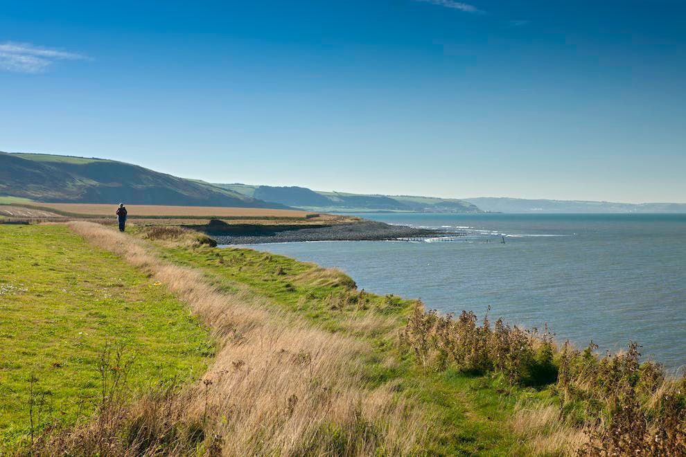A coastal pathway runs alongside the water, leading towards rolling hills in the distance. Lush green grass and wildflowers line the path, while calm waves ripple against the shore under a clear blue sky.