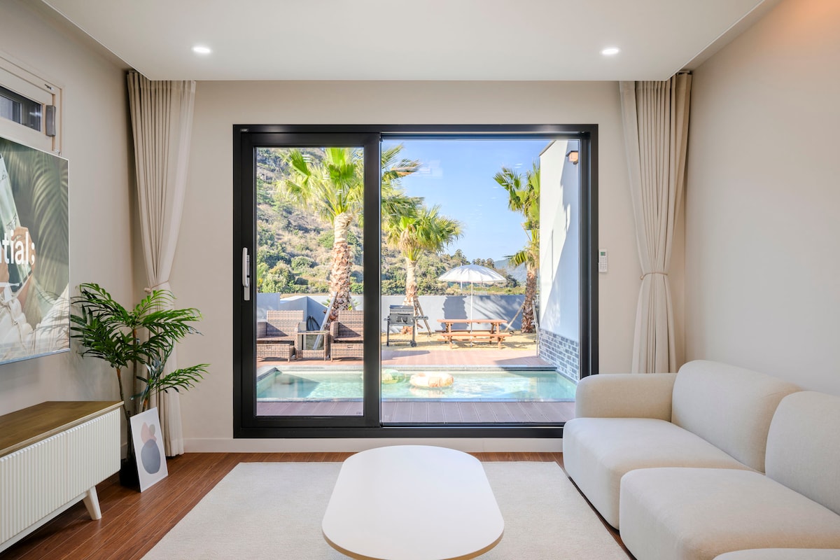 A spacious living area features large glass doors that open to an outdoor terrace adorned with palm trees and a swimming pool. A comfortable sofa sits next to a minimalist coffee table, inviting relaxation and enjoyment of the scenic view.