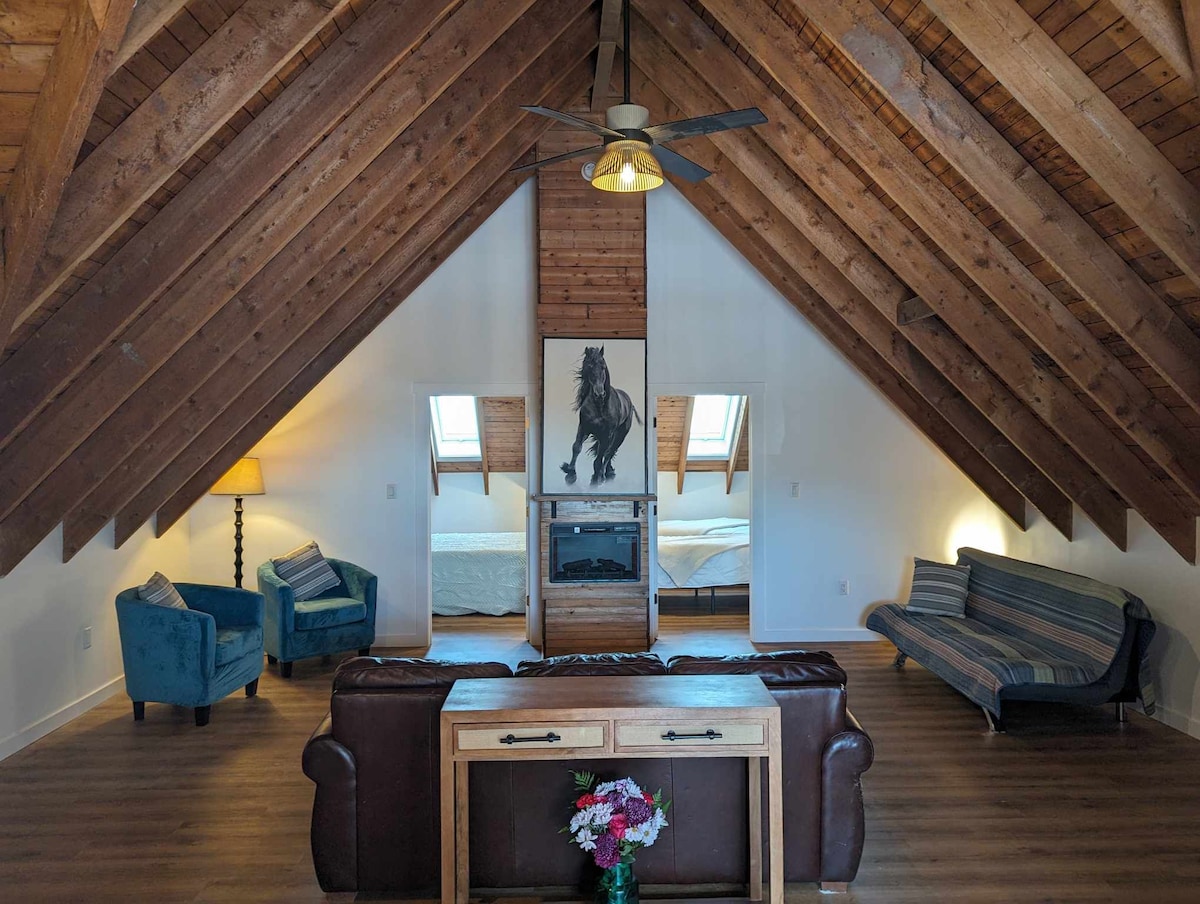 A spacious living area features a combination of comfortable seating arrangements. Vaulted wooden ceilings with exposed beams enhance the overall charm. Natural light fills the space through skylights, highlighting a decorative horse painting above an electric fireplace. A wooden table with flowers adds a touch of warmth.