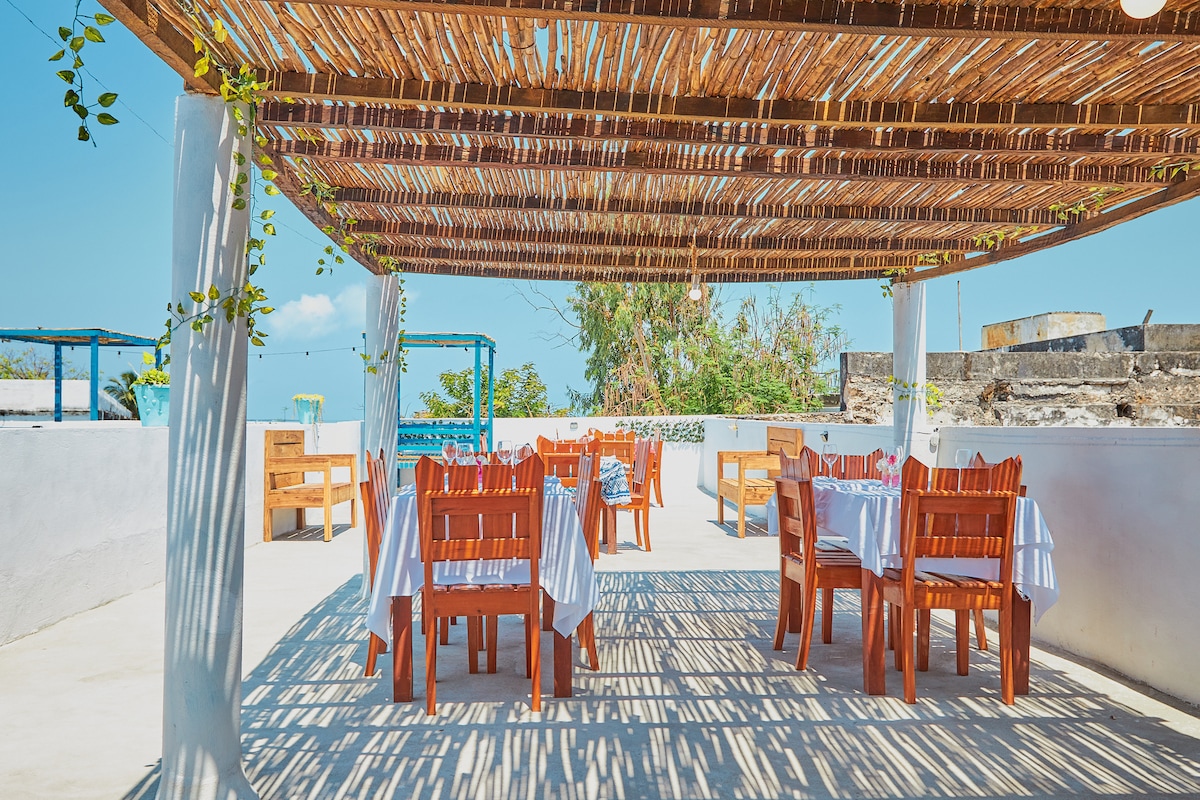 An outdoor dining space is presented with several wooden tables set under a bamboo canopy. Natural light creates playful shadow patterns on the white surface. Surrounding greenery adds a touch of nature to the inviting, open environment.