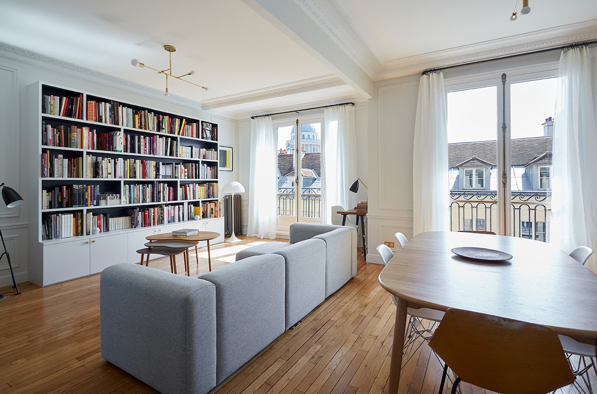 A bright living area is presented, featuring a modern gray couch facing a large bookshelf filled with neatly arranged books. A round dining table with light wooden chairs is situated nearby. Floor-to-ceiling windows allow natural light to fill the space, with soft curtains gracefully framing the views.