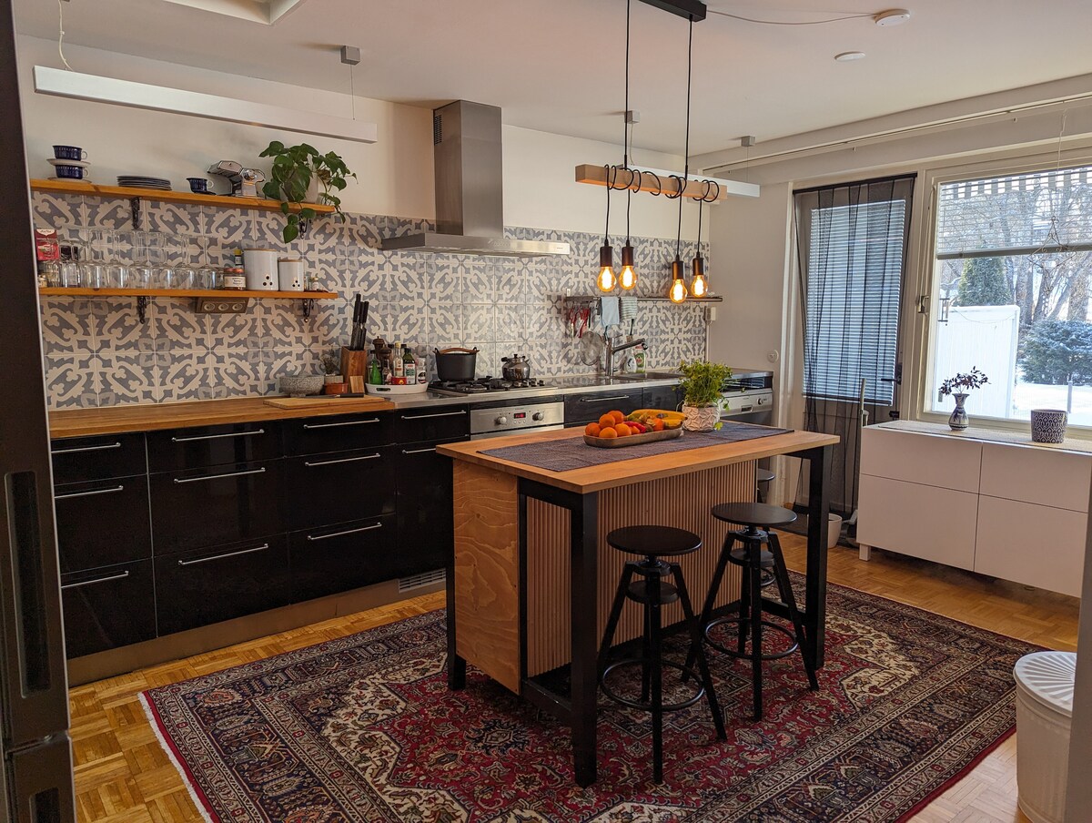 A modern kitchen is featured with dark cabinetry and patterned backsplash. A central wooden island with seating for two is highlighted. Warm lighting from pendant fixtures creates a welcoming space. Large windows allow natural light, and a decorative rug adds a touch of comfort.